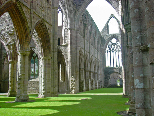 Photograph of the nave and stonework of Tintern Abbey, now in ruins, but with tall walls still standing with arched side chapels and spaces. The ground is a beautiful flat carpet of green grass.
