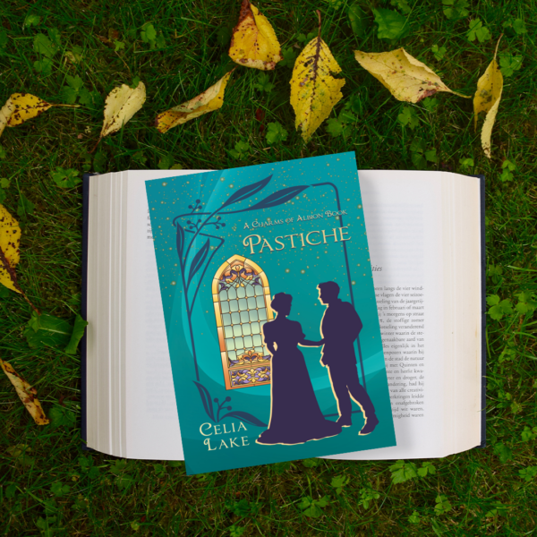 Copy of Pastiche on an open book on grass with yellow leaves. The cover has a silhouetted man and woman in Edwardian dress, in front of a golden stained glass window on a deep teal background.