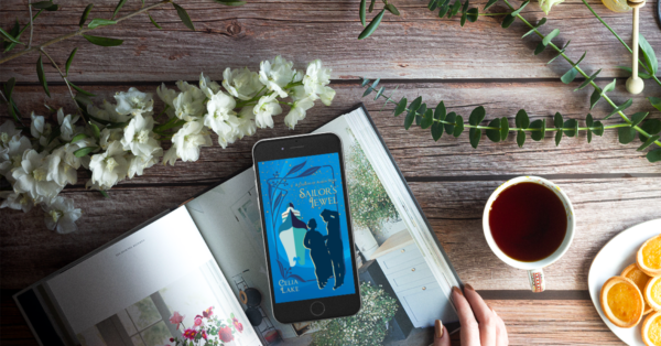 Cover of Sailor's Jewel displayed on a phone, resting on a desk with flowers, eucalyptus boughs, and a book about a garden.