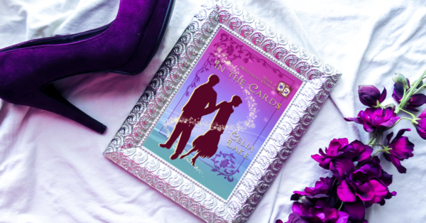 Copy of In The Cards displayed in a silver picture frame, with a deep purple high heel shoioe and orchids framing the image. The cover has a woman turning away from a man in silhouette, in shades of vivid pinks and purples. 