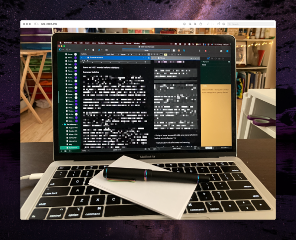 A laptop with a black fountain pen with shimmery metal lying on the keyboard on top of some index cards. The screen shows a Scrivener window with multiple areas of text and notes, in shades of dark green, black, and gold backgrounds. The content of the screen is described in the following text.