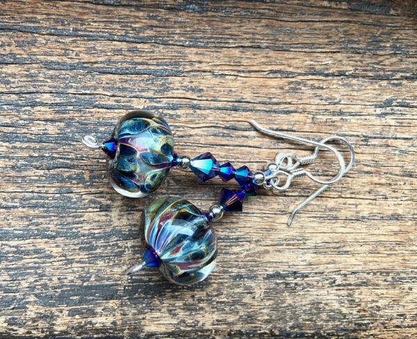A pair of earrings shimmering with blue irridescence lie on a wooden table. Two smaller blue-green beads lead into a larger one, with a larger lampwork glass bead at the bottom swirling with peacock colours of blues, greens, and purples as well as clear glass. 