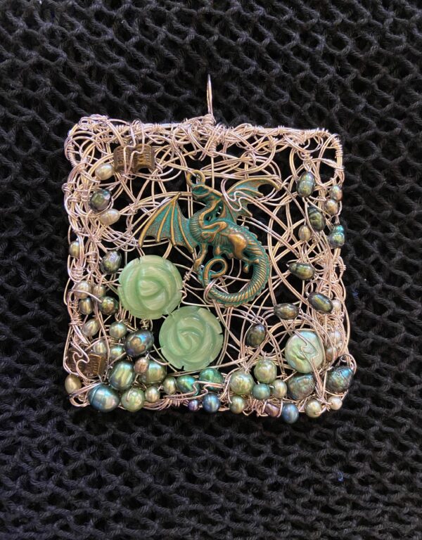 A square pendant is woven thickly with silver wire, forming the backdrop for a jade-green and gopper dragon charm, pale green glass rose beads, and droplets of shimmering pearls in the pale colours of the water: opalescent green, blue, and white. 