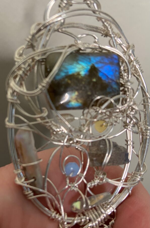 The focus on this pendant is fuzzy, in order to capture the flash of the labradorite that is the main stone of the piece. The blue flash lights up the top of the bead, with a black shape forming the outline of a house with a peaked roof. Smaller labradorite beads, swirled around with silver wire, make up the rest of the pendant. 