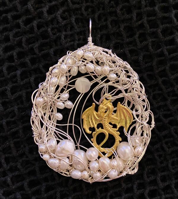 A circular pendant of intricate woven silver wirework forms the frame for a golden dragon charm. A single transluscent white bead hangs in the air above the dragon, while layers of pearls of different sizes frame the ground and the sky around the two.