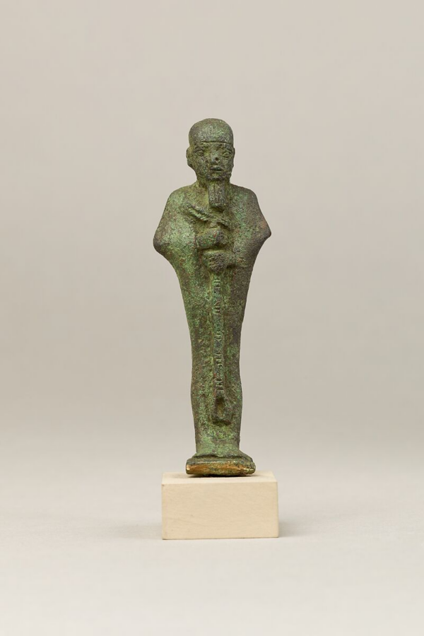 A metal statue of the god Ptah, with somewhat unbalanced facial features and unevenly carved eyes, wearing the tight-fitting cap, straight beard, and carrying a scepter. 