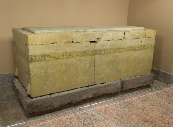 A massive limestone sarcophagus on a stone dais, with inscriptions running in a strip around the outside, about a hands-breadth down from the top of the base. They were first painted green, then outlined in black, creating delicate details, with the rest of the sarcophagus left the natural colour of limestone. 